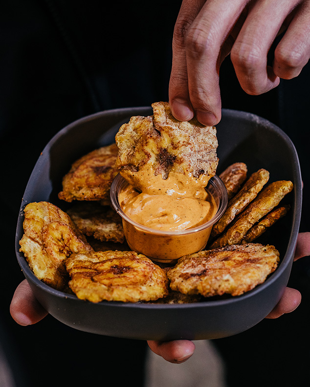 A hand dips a plantain chip into a sauce within a bowl filled with more chips.
