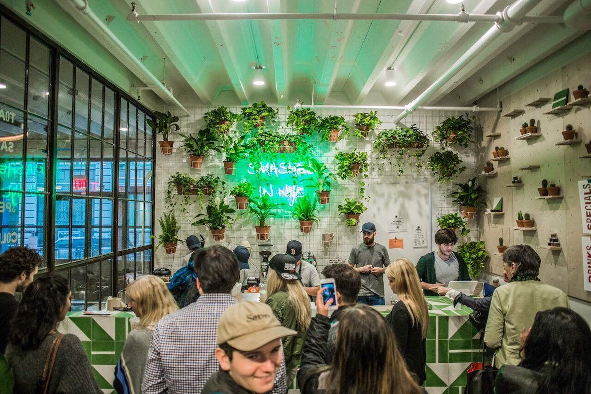 People wait in line at a restaurant counter. Behind the counter there is a green neon sign surrounded by plants.