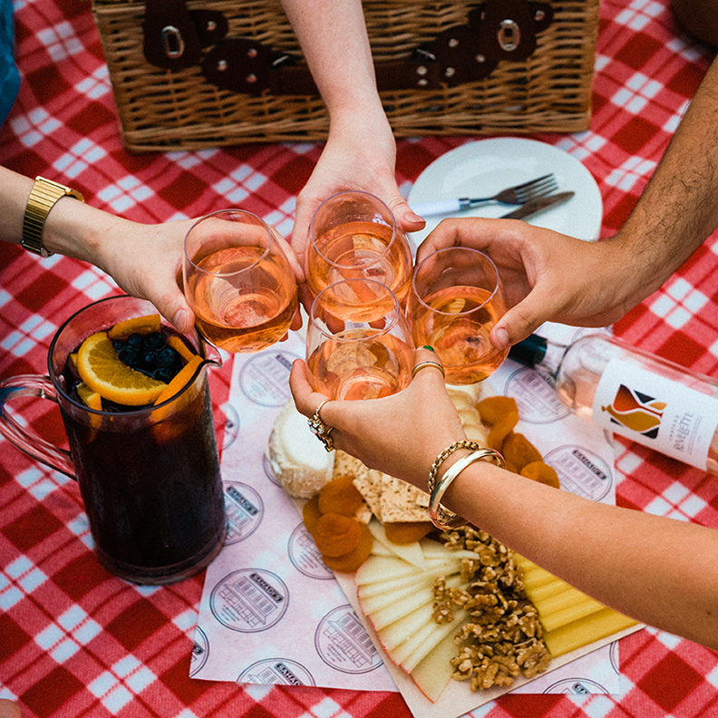 People grab wine glasses off of a picnic blanket. Food and items are on the blanket.