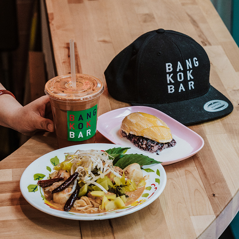 A Bangkok Bar hat, two plates of food, and a iced drink are on a wood counter.