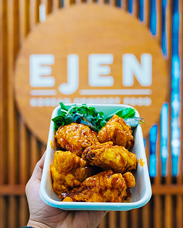 A hand holds up a plate of food in front of a sign for Ejen.
