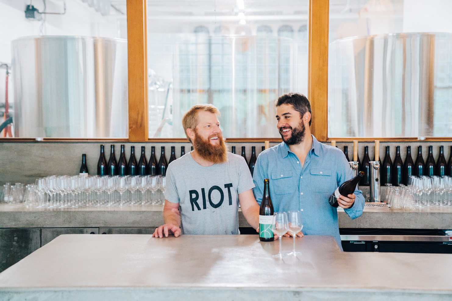 Two people are holding bottles of wine behind a bar.