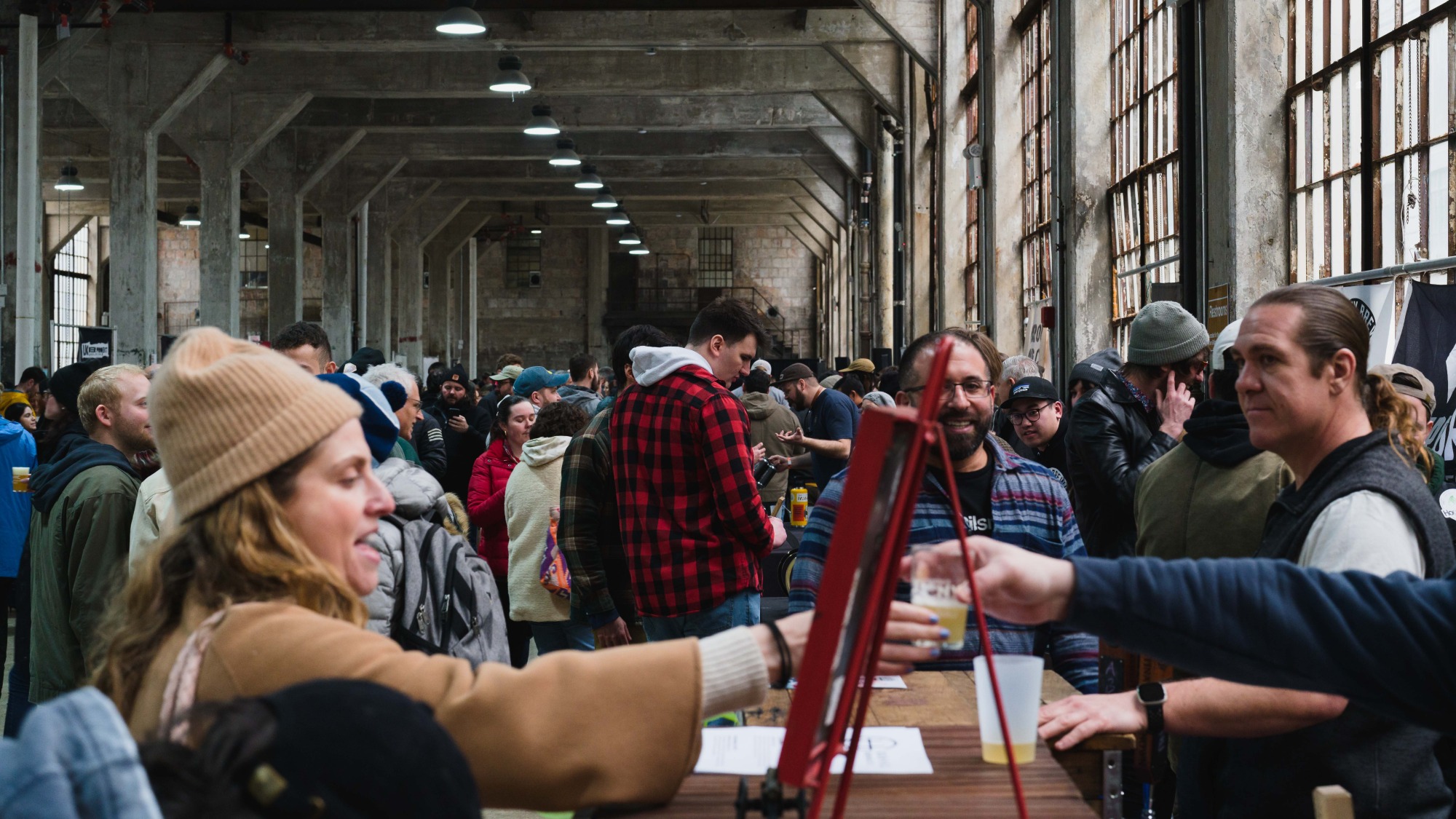 A crowd of people in a large industrial space. People hold beers.